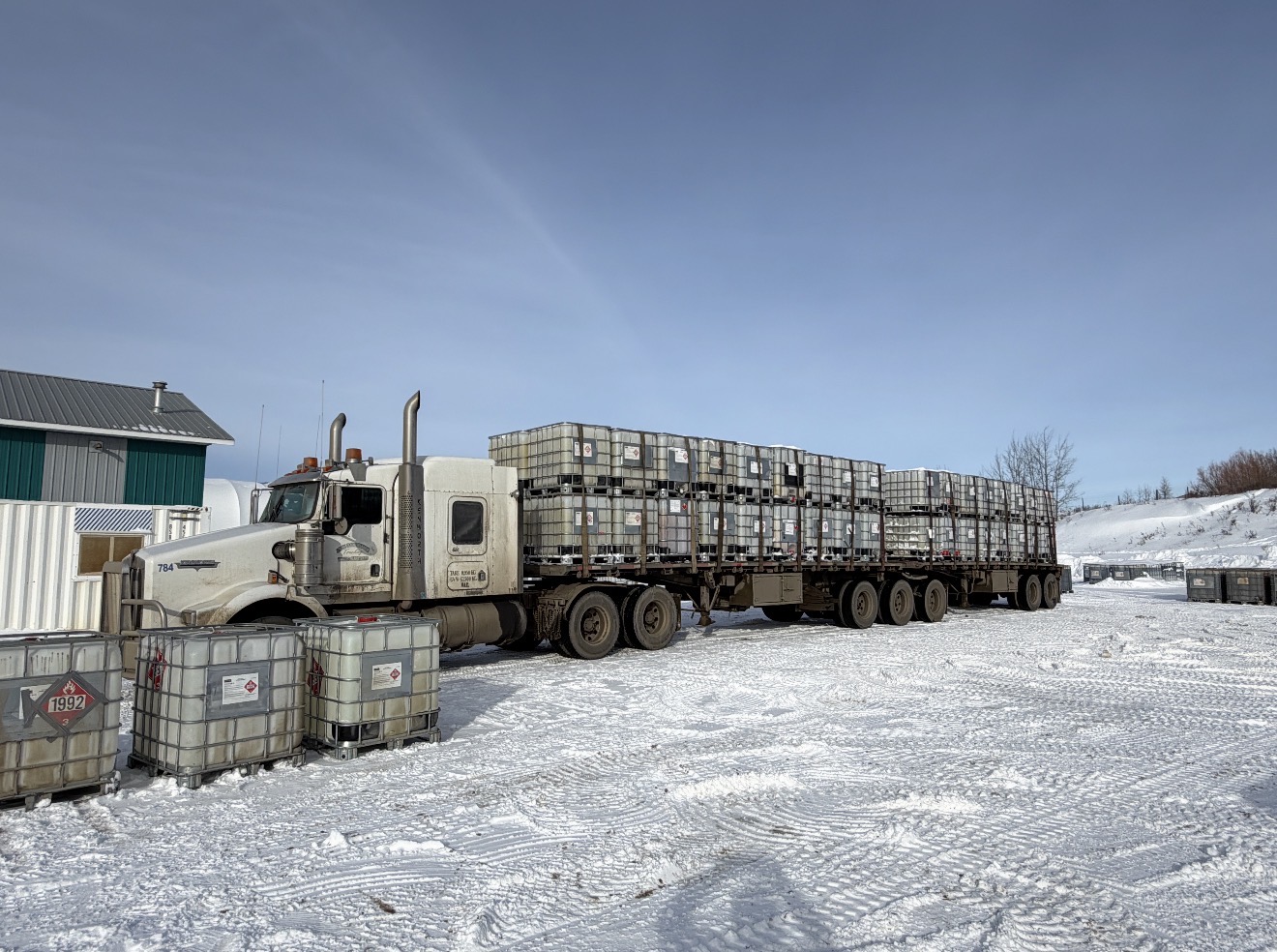 Kenworth tractor with flatbed loaded with placarded IBC totes at a winter site